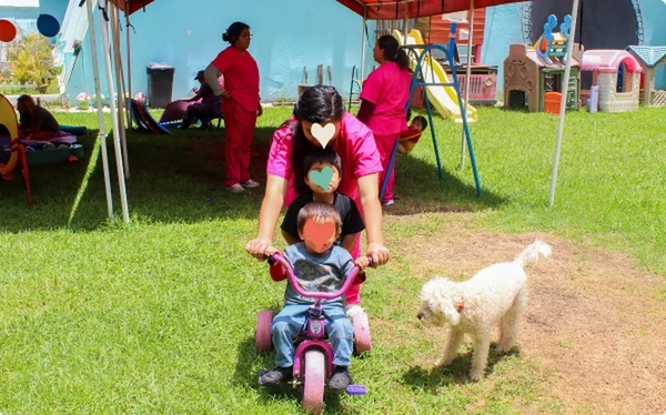 Cuidadora acompañando a un niño en triciclo por el jardín, junto a un perrito blanco