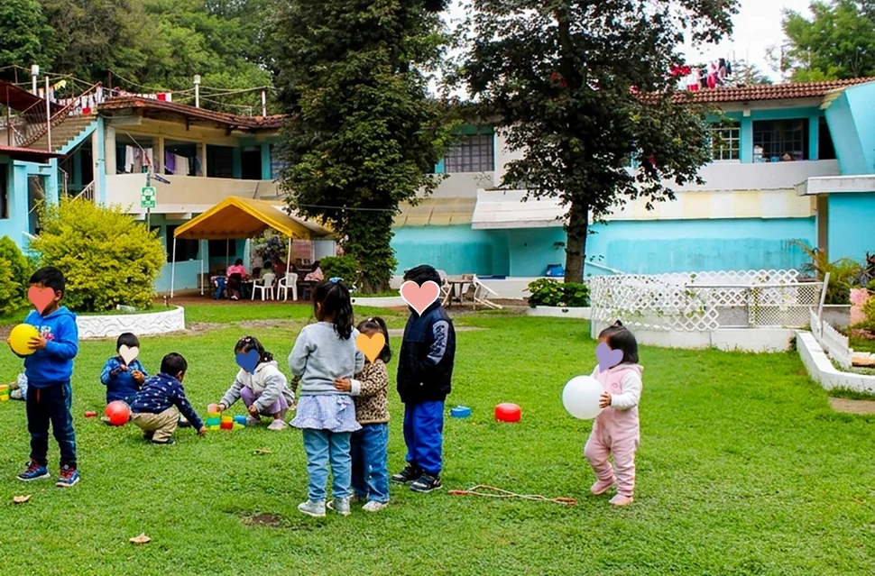 Niñas y niños jugando con pelotas en el jardín frente a la casa del Hogar, con los rostros cubiertos por corazones